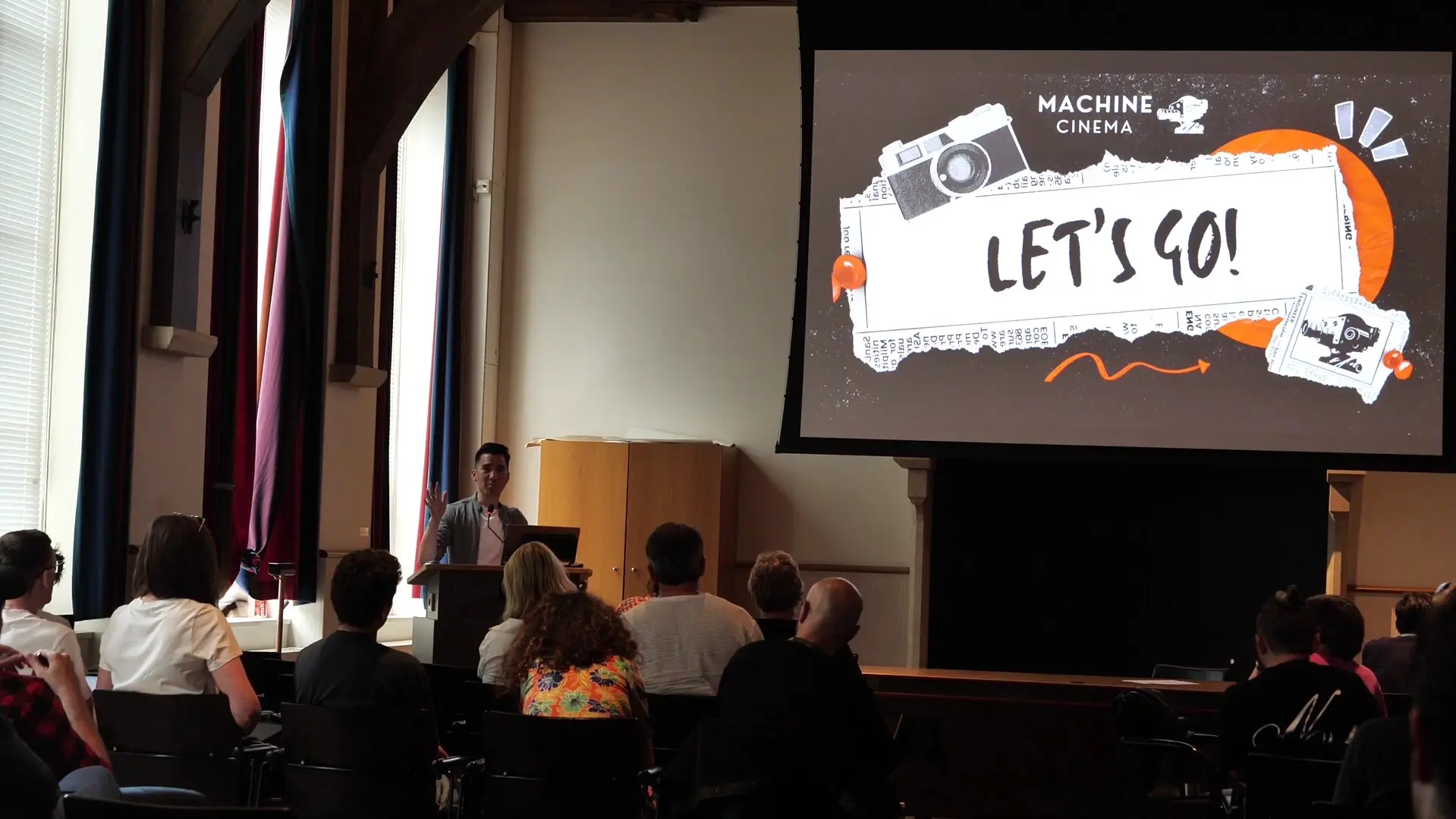 Machine Cinema host on stage with a 'Let's Go!' slide — the kind of room MosaicJam plays into.